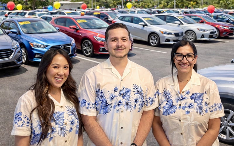 Lo-Ann H., Hector L., and Laura V. standing in a dealership parking lot.