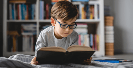 Young boy with glasses leaning on his bed reading a book