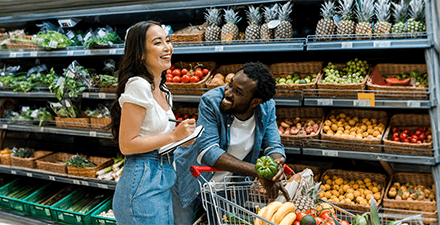 happy african american man looking at asian woman with notebook near shopping cart with groceries