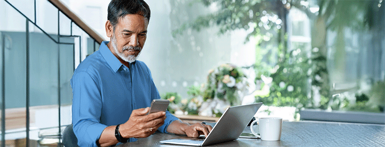 Smiling mature man with white, grey stylish short beard using smartphone and laptop to view his banking information.